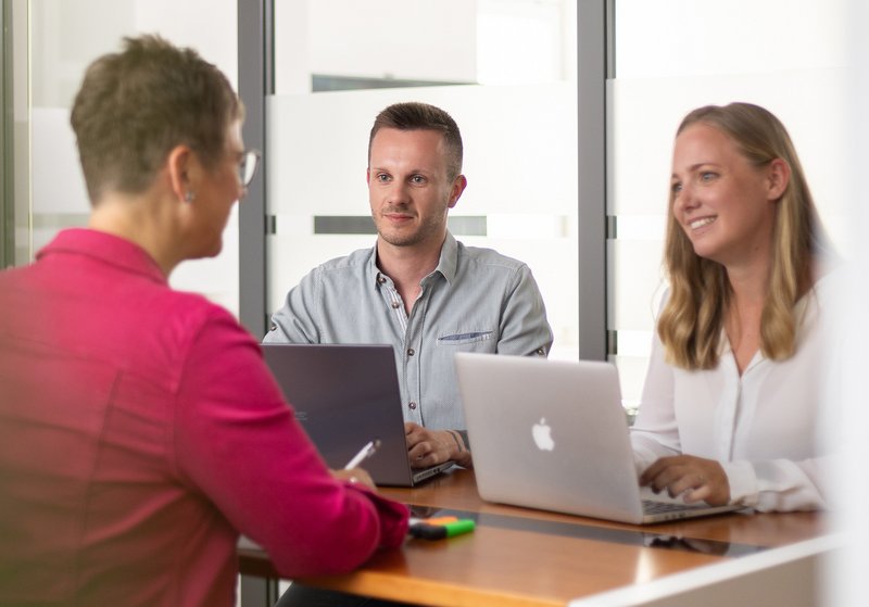 Drei Mitarbeitende sitzen am Tisch in einem Meeting, sprechen miteinander und arbeiten am Laptop