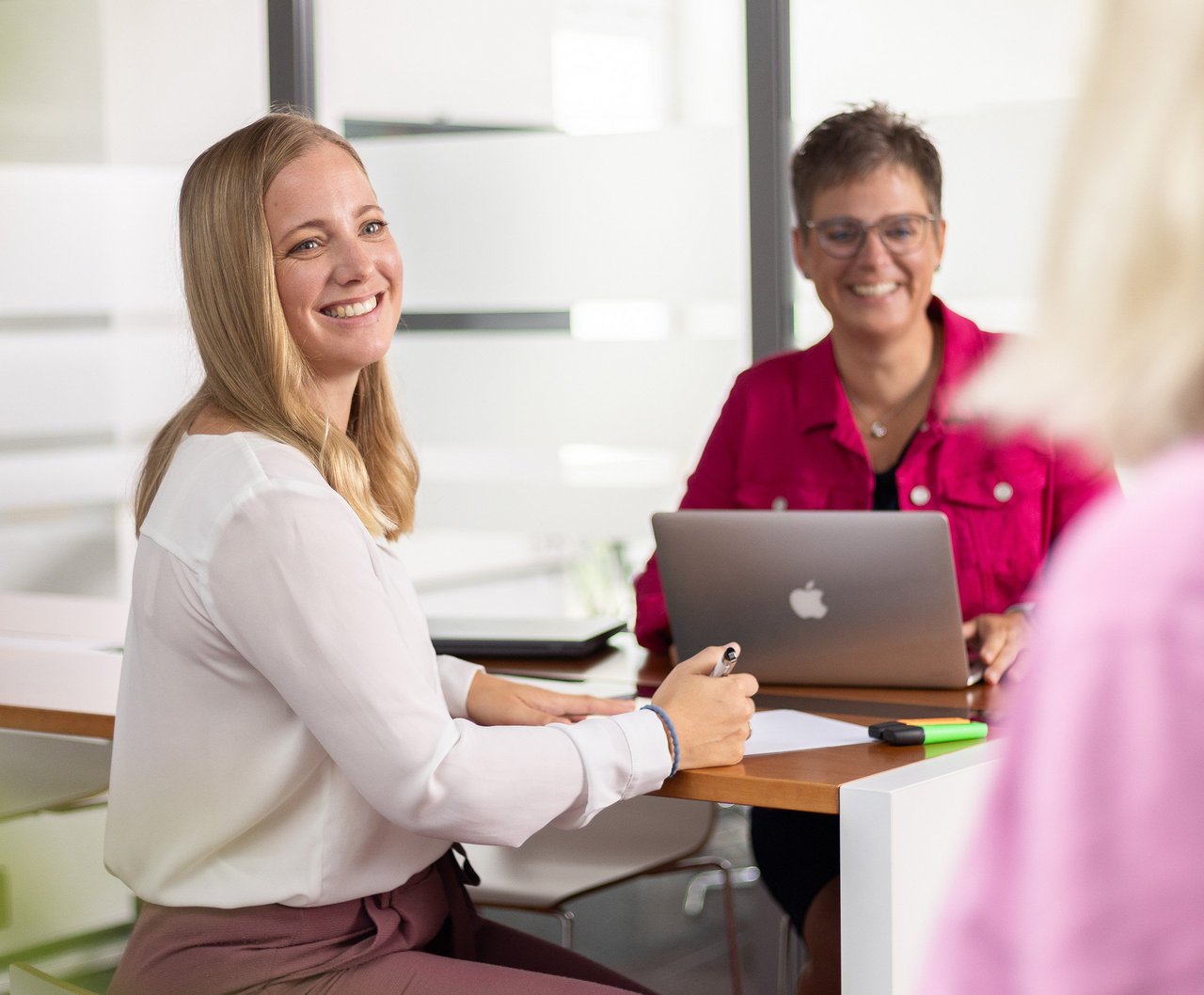 Zwei Mitarbeiterinnen der Insolvenzkanzlei sitzen an einem Besprechungstisch und lächeln in die Kamera